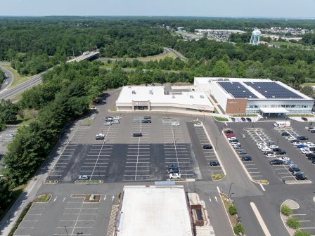 An aerial view of a large parking lot with few cars, surrounded by greenery and buildings, under a clear sky.