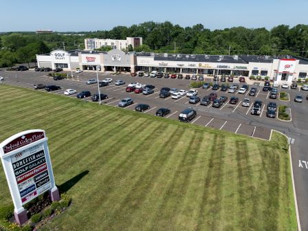 A shopping center with multiple stores, a parking lot with cars, and a sign listing businesses, surrounded by grass and trees.