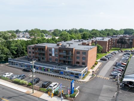 The image shows a brick apartment building with shops at the ground level, surrounded by parked cars and greenery on a clear day.