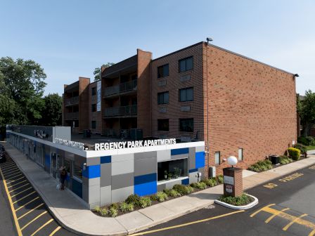 The image shows a brick building with a sign reading "Regency Park Apartments," surrounded by landscaping and parking spaces.