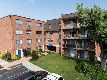 The image shows a red-brick apartment building with a small parking area, green lawn, and trees, set under a clear blue sky.