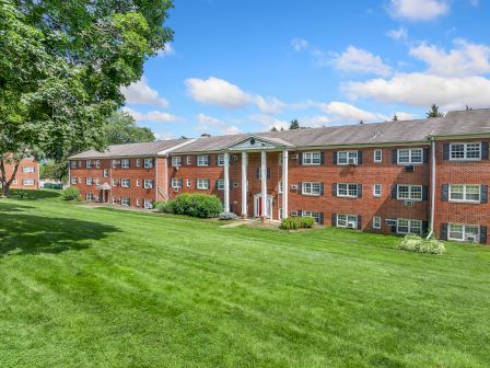 A large, red-brick apartment building with white columns surrounded by a grassy lawn and trees under a blue sky with clouds.
