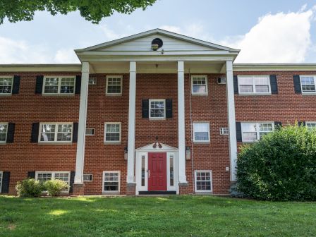 A red brick building with a red door, white columns, and black shutters, surrounded by greenery and a clear blue sky.