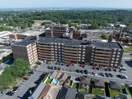 An aerial view of a large, rectangular building with parking lot, surrounded by trees and residential homes.