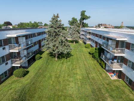Two apartment buildings face each other with a grassy courtyard and trees in between, under a clear blue sky.