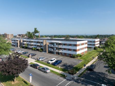 The image shows a three-story apartment building with a parking lot, surrounded by trees and a street intersection in the foreground.