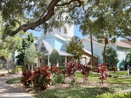 The image shows a light blue building surrounded by lush greenery and red plants, with a winding path in the foreground.