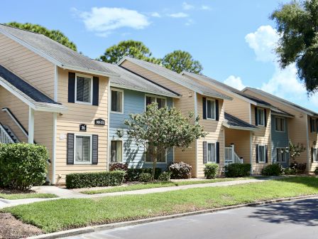 A row of light-colored townhouses with small gardens and trees, set against a clear sky, are shown.