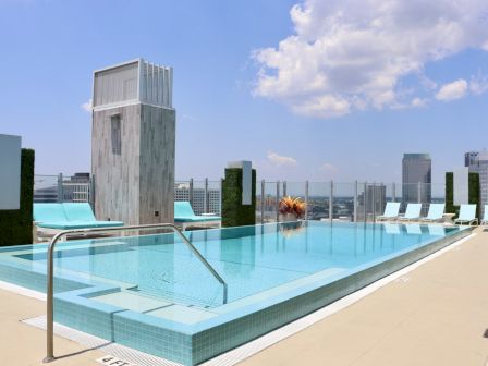 A rooftop infinity pool with lounge chairs, city skyline, and clear sky in the background.