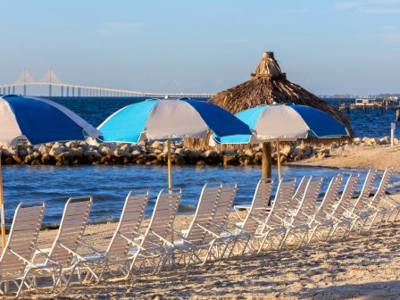 Beach scene with rows of empty lounge chairs, blue and white umbrellas, a thatched hut, and a bridge in the background over the water.