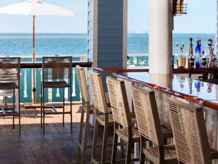 Outdoor bar with high stools, wooden deck, and ocean view under a sunny sky, featuring a row of liquor bottles on the counter.
