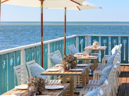 Outdoor seaside dining area with neatly set tables, white chairs, and umbrellas, offering a scenic ocean view for a relaxing experience.