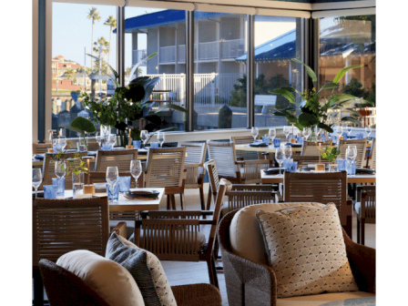 A stylish restaurant interior with wicker chairs, plush cushions, chandeliers, and large windows letting in natural light.