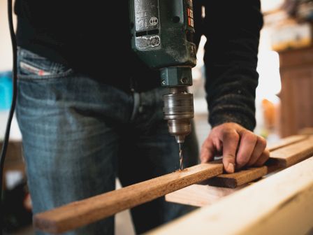 A person is using an electric drill to bore a hole into a wooden plank in a workshop setting.
