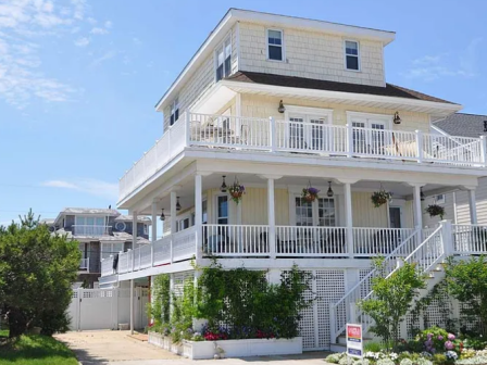 Three-story light yellow house with wraparound white porch, hanging plants, and a manicured garden under a sunny blue sky.