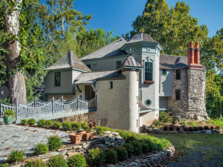A charming gray-and-stone cottage with turret, chimney, stone path, and lush landscaping in a sunny garden setting.