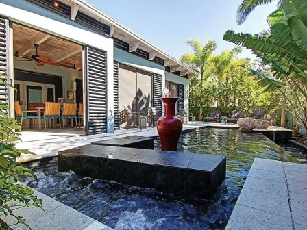 A modern house with an outdoor pool and water feature; a red vase sits on a black platform, tropical plants surround a sunny patio.
