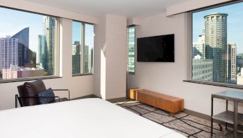 A modern room with a bed, chair, TV, and large windows offering a city skyline view.