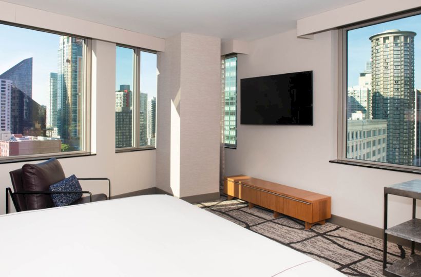 A modern room with a bed, chair, TV, and large windows offering a city skyline view.