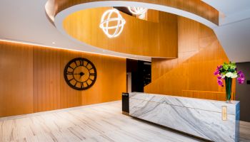 A modern lobby with a marble reception desk, wooden walls, large clock, spiral staircase, and a flower arrangement on the counter.