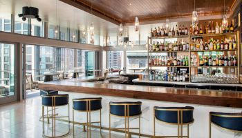 The image shows a modern bar with a wooden countertop, bar stools, and shelves of bottles, set in a room with large windows.