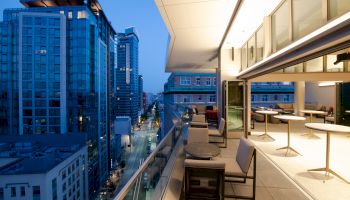 A modern balcony overlooking a cityscape at dusk, with tables, chairs, and buildings lit up.