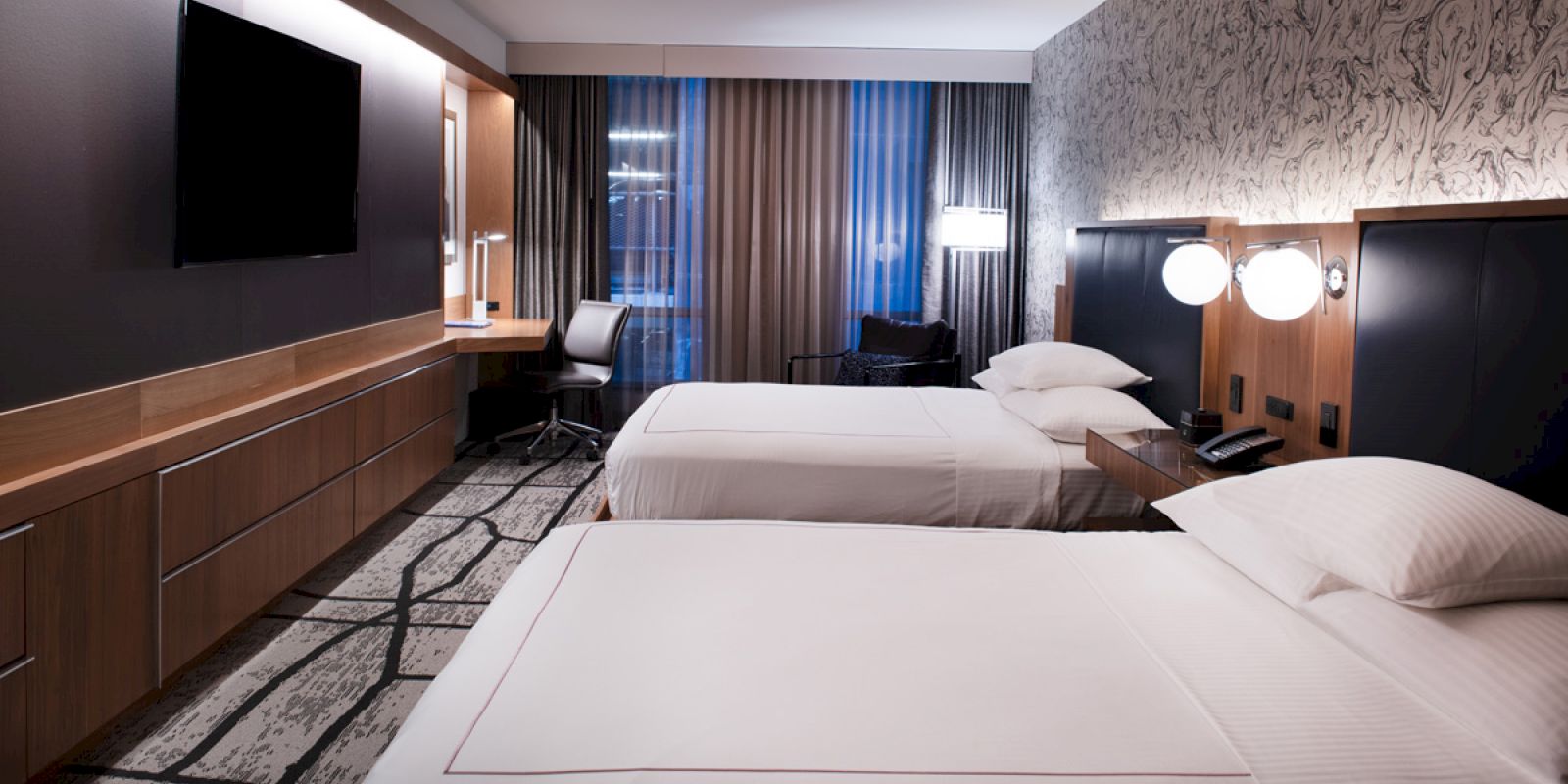 The image shows a modern hotel room with two beds, a desk, a flat-screen TV, and patterned carpet and wallpaper.