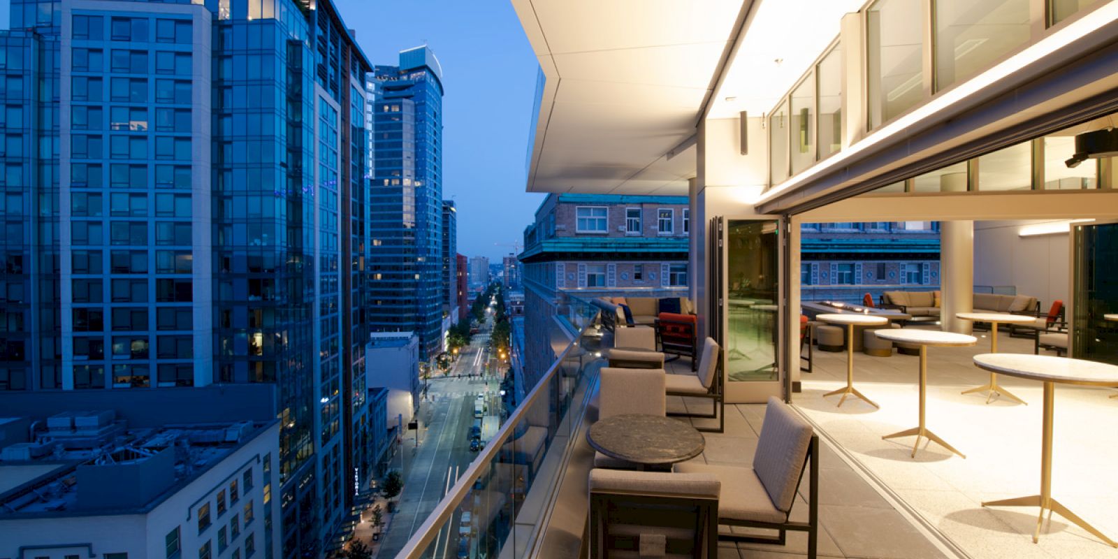A modern city view from a balcony at dusk, with buildings and street lights on one side and outdoor seating on the balcony.