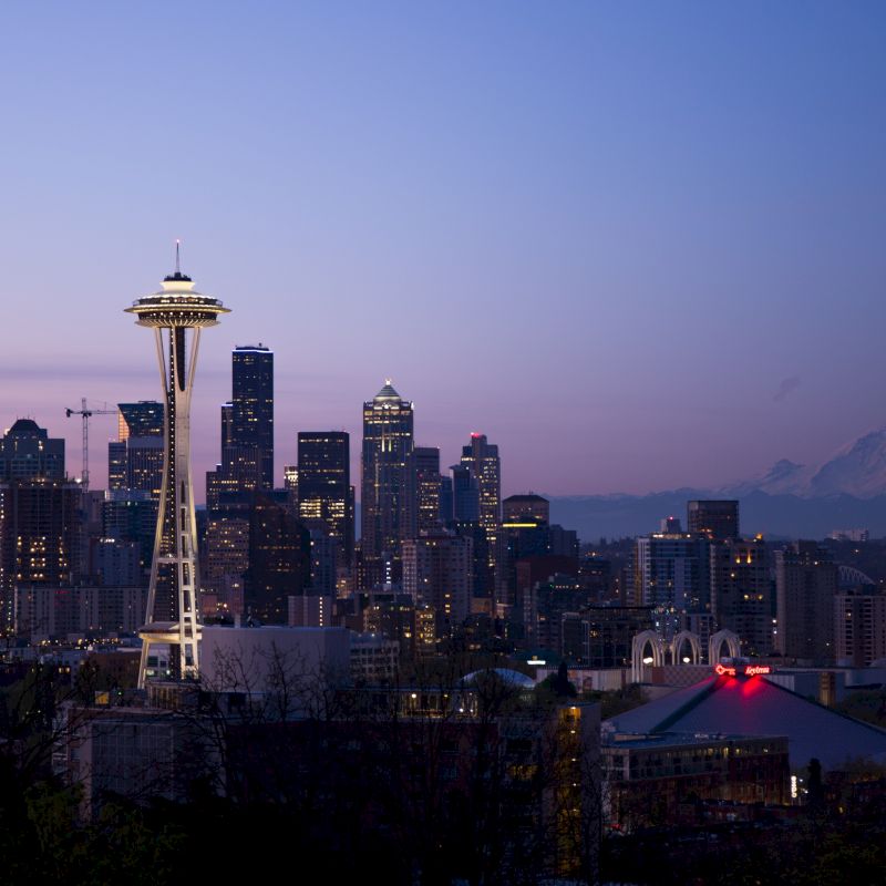 Seattle skyline at dusk, featuring the Space Needle and Mount Rainier in the background, against a clear blue sky.