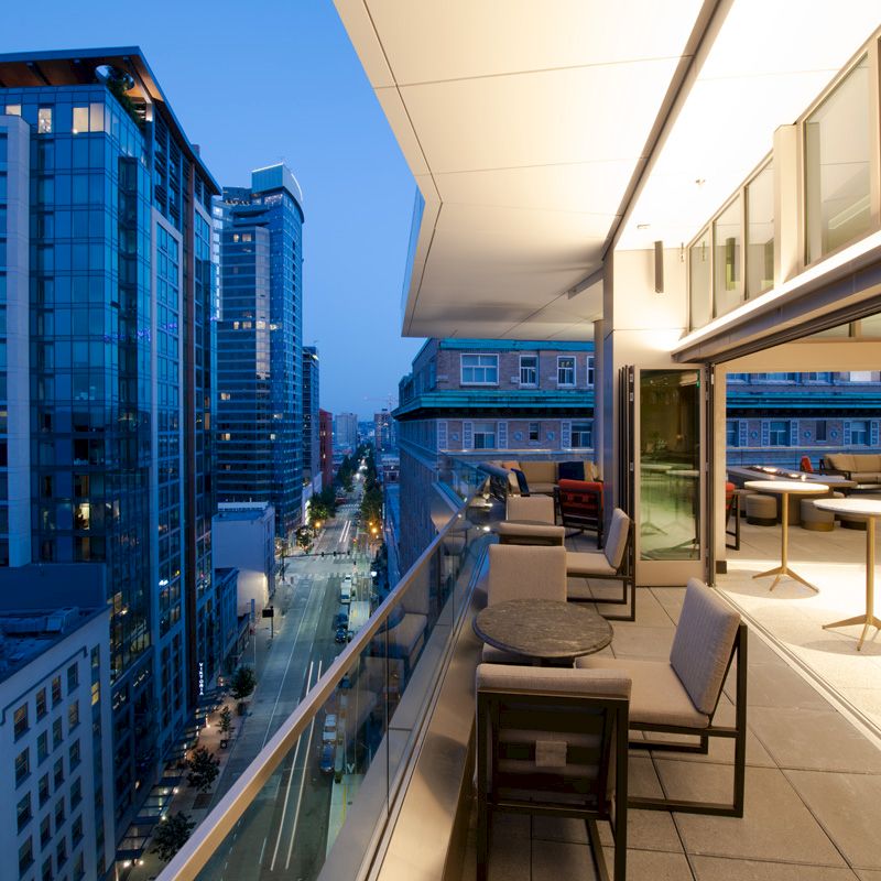 A modern cityscape at dusk, viewed from a rooftop terrace with tables and chairs, overlooking tall buildings and a street below.