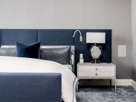 A modern bedroom features a blue upholstered bed, a white nightstand with a lamp, and decorative elements on a textured gray wall.