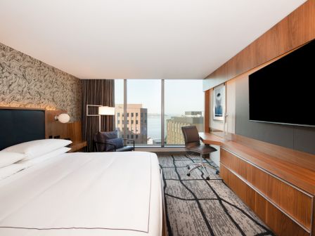 A modern hotel room with a large bed, desk, chair, and television, featuring a cityscape view through a wide window.