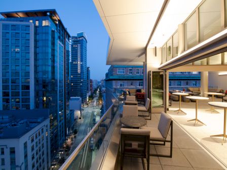 A modern rooftop terrace at dusk with city buildings visible; features seating and tables along the glass railing.