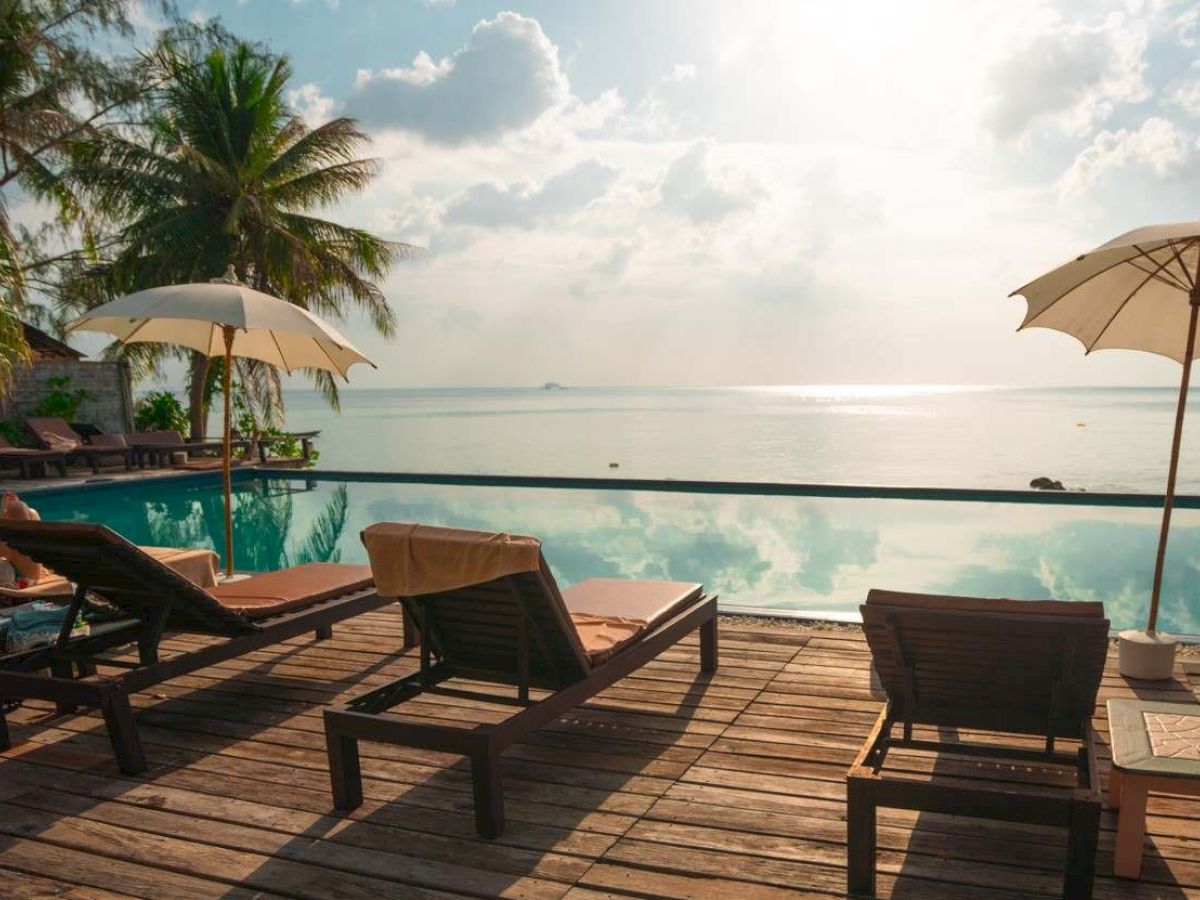 Lounge chairs and umbrellas overlook an infinity pool with an ocean view, surrounded by palm trees under a partly cloudy sky.