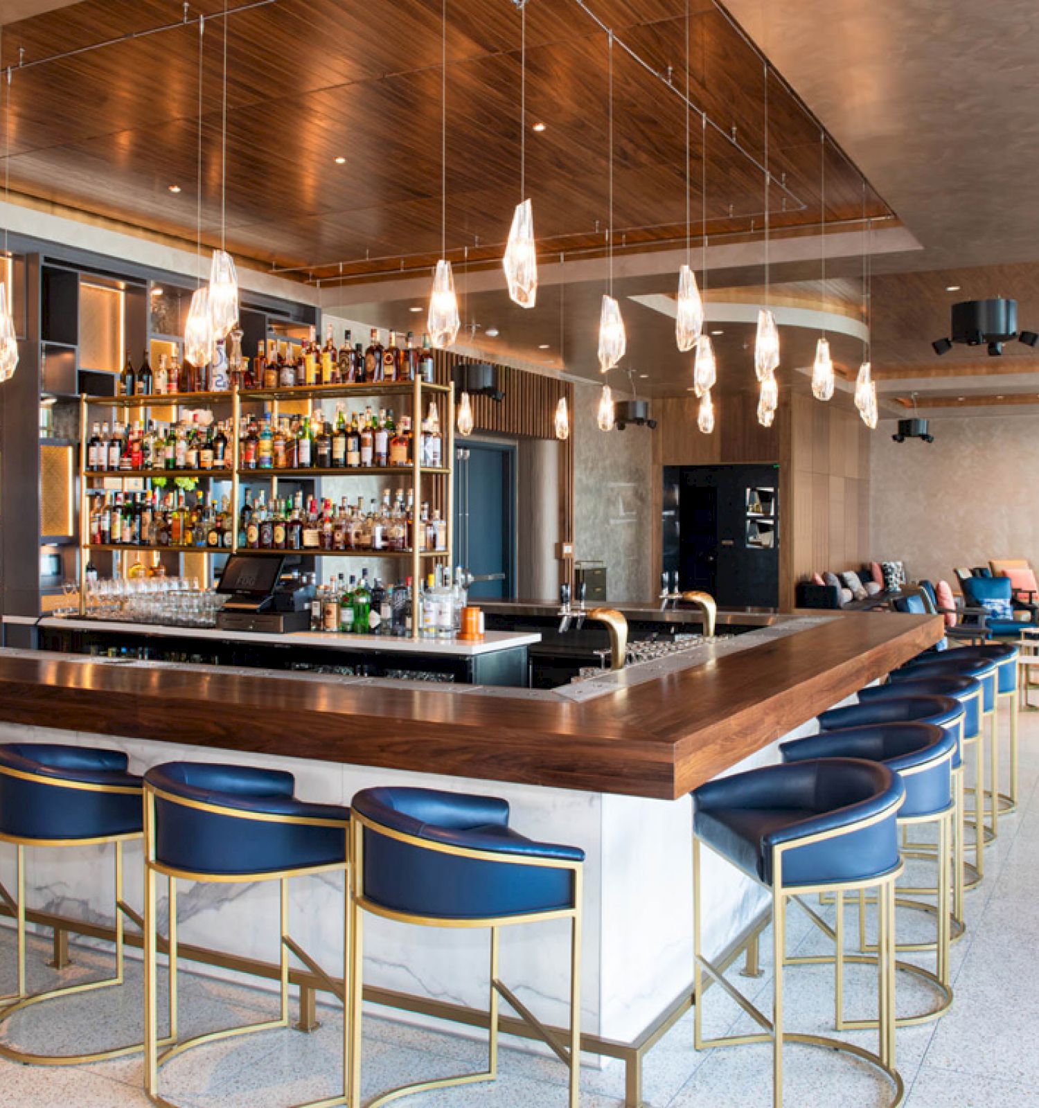 A stylish bar with modern lighting, a well-stocked shelf, blue chairs, and a cozy seating area in the background.