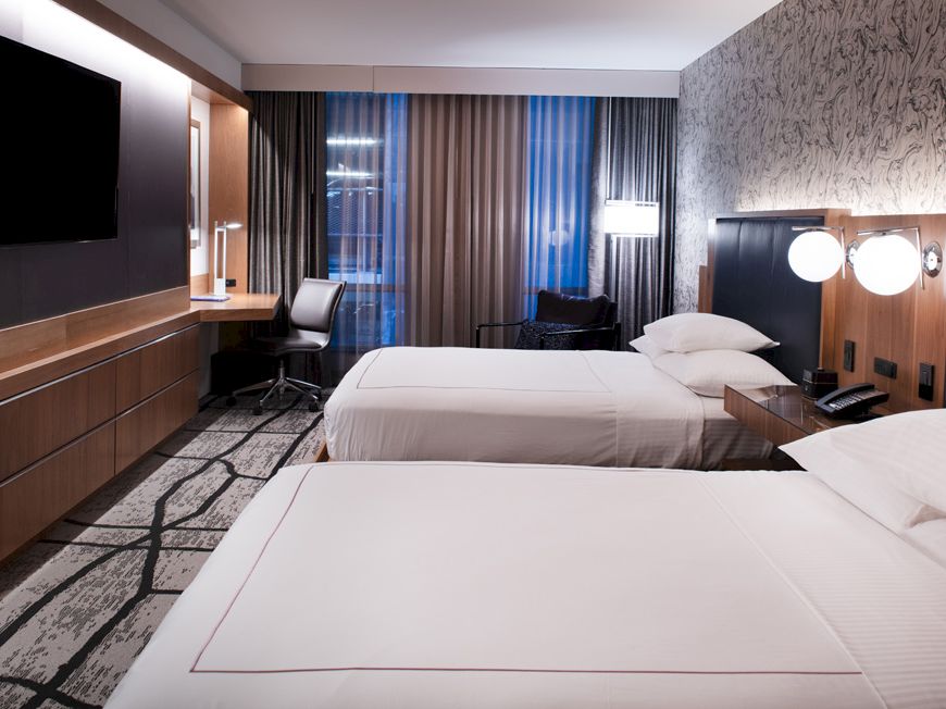 The image shows a modern hotel room with two beds, a desk, chair, lamps, a wall-mounted TV, patterned carpet, and curtains.
