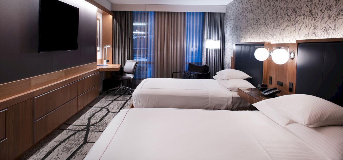 The image shows a modern hotel room with two beds, a desk, chair, lamps, a wall-mounted TV, patterned carpet, and curtains.