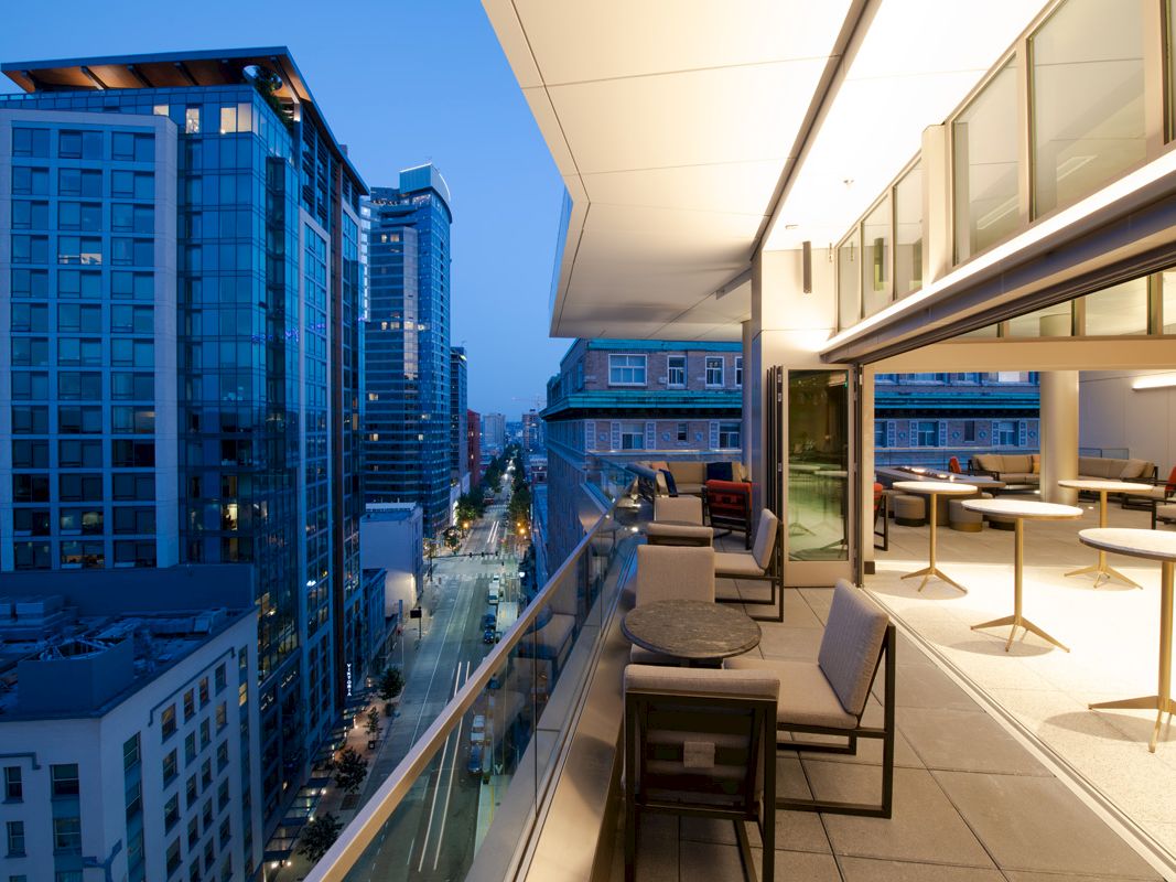 A modern cityscape at dusk showing a rooftop terrace with seating, overlooking tall buildings and a lit street below.