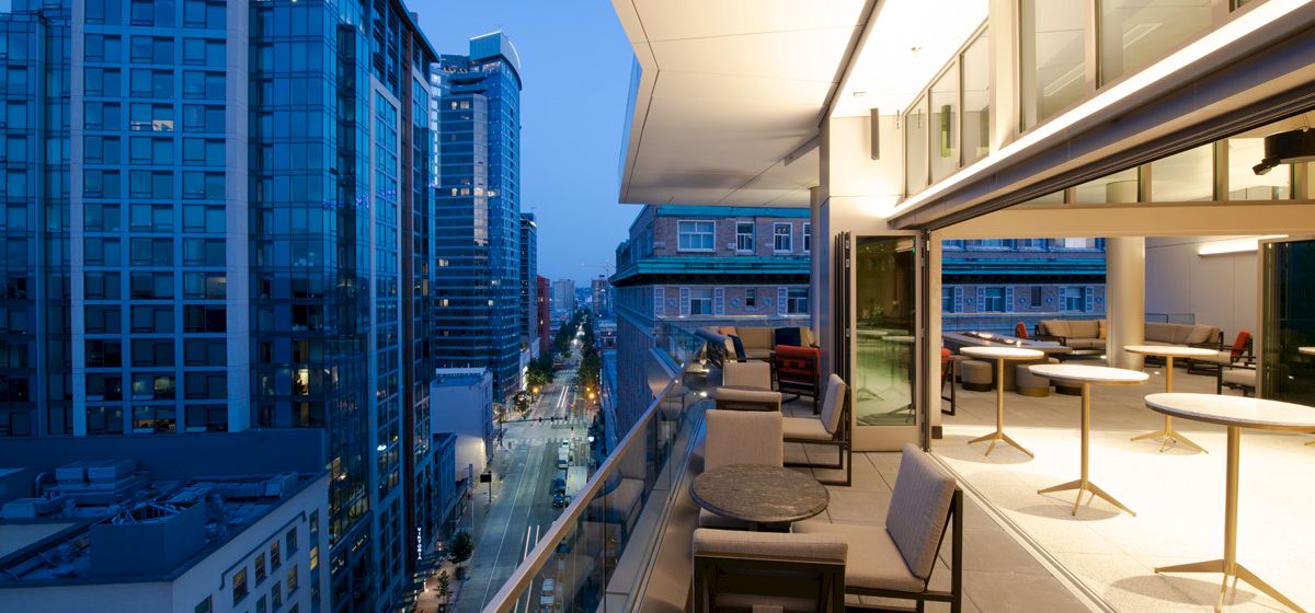 A modern cityscape at dusk showing a rooftop terrace with seating, overlooking tall buildings and a lit street below.