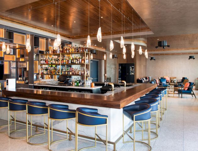 A modern bar with blue stools, wooden accents, and hanging lights, featuring a well-stocked drink shelf and seating area in the background.
