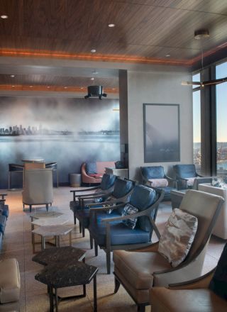 A well-lit lounge area with comfortable seating, large windows showing a waterfront view, and a wall mural depicting a city skyline.