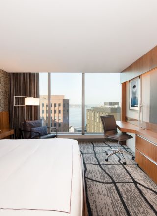 A modern hotel room with a large bed, desk, chair, and TV, overlooking a cityscape through a wide window.
