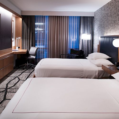 The image shows a modern hotel room with two beds, a TV, desk, chair, and stylish decor, featuring patterned carpet and wallpaper.