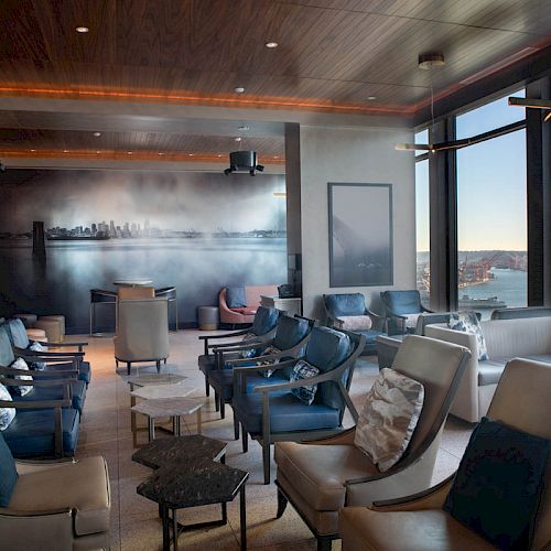 A modern lounge with cushioned chairs, large windows offering a scenic view, and ambient lighting, featuring a wall mural of a city skyline.
