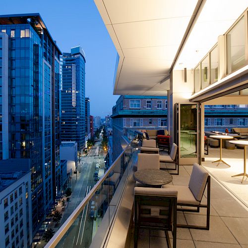 A modern rooftop patio with seating overlooks a cityscape at dusk, featuring tall buildings and illuminated streets.