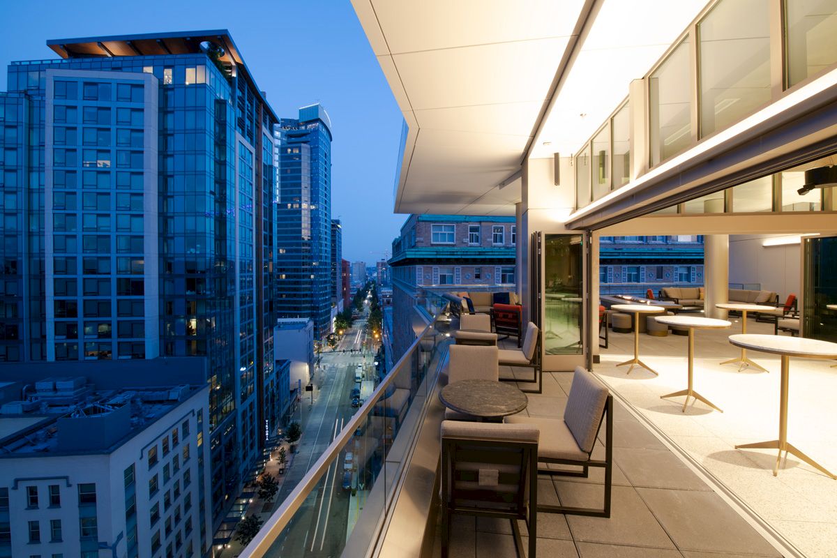Modern urban rooftop patio with tables and chairs overlooking a cityscape of tall buildings at dusk.