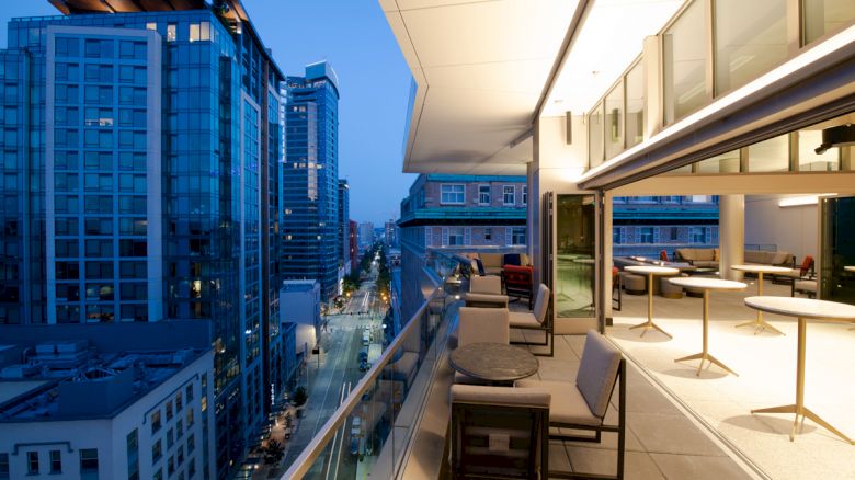 Modern urban rooftop patio with tables and chairs overlooking a cityscape of tall buildings at dusk.