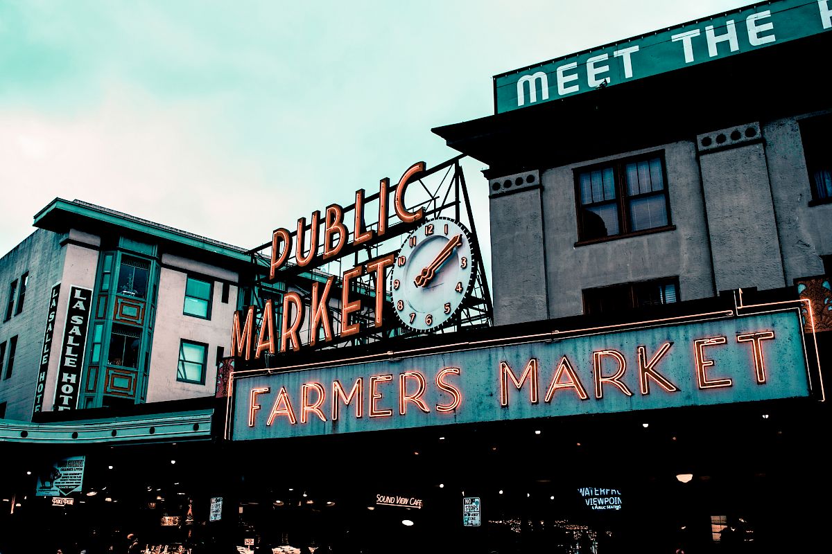 The image shows a public market with neon signs reading "PUBLIC MARKET" and "FARMERS MARKET," along with a clock and surrounding buildings.