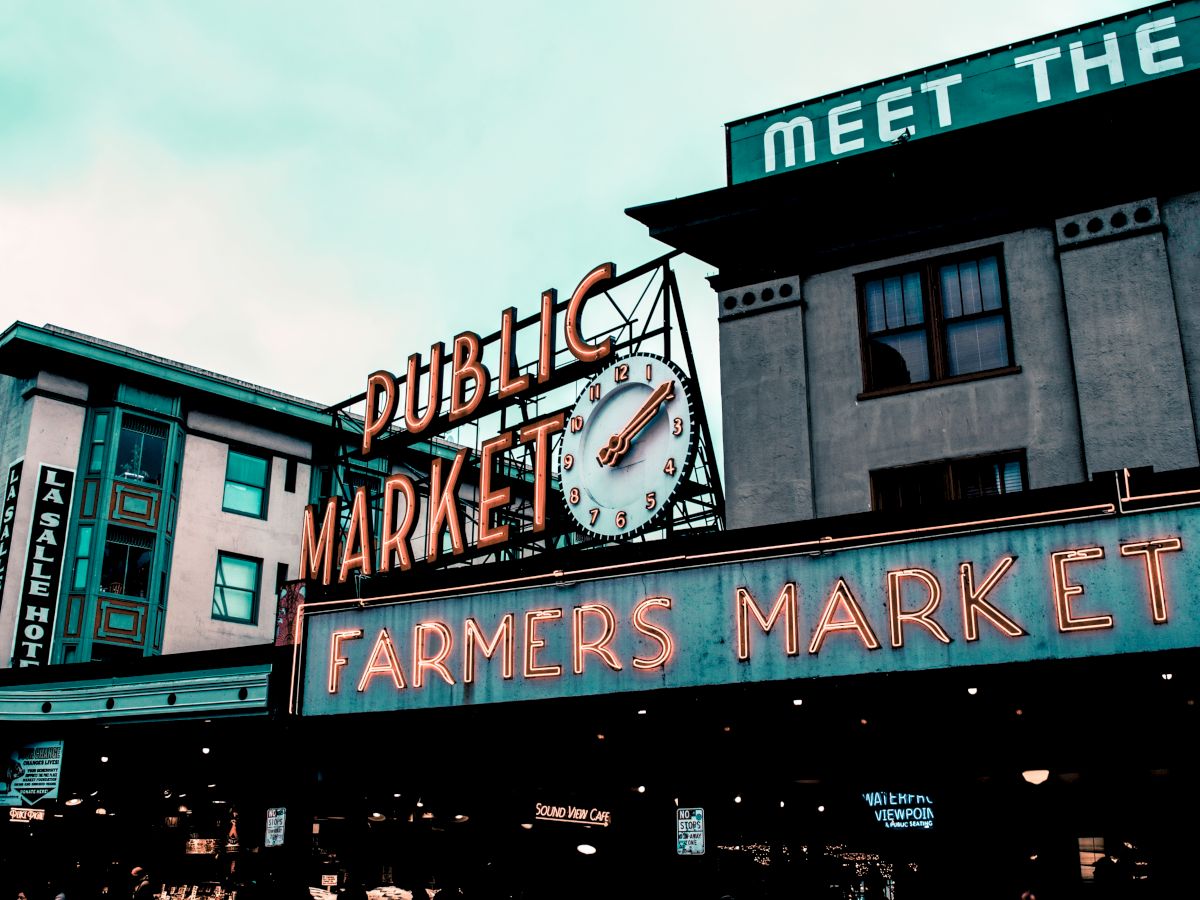 The image shows a public market with neon signs reading "PUBLIC MARKET" and "FARMERS MARKET," along with a clock and surrounding buildings.