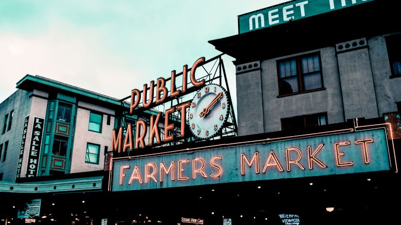The image shows a public market with neon signs reading "PUBLIC MARKET" and "FARMERS MARKET," along with a clock and surrounding buildings.